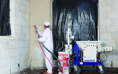 Pracownik w białym kombinezonie natryskuje tynk na ścianę z bloczków. Obok stoi maszyna m-tec M300 i worek z napisem "lekkie tynk".
