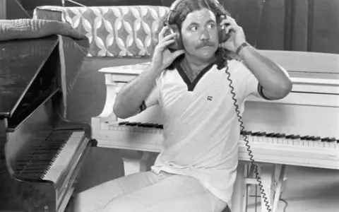 Billy Powell hört mit Kopfhörern Musik, während er neben einem Klavier sitzt.