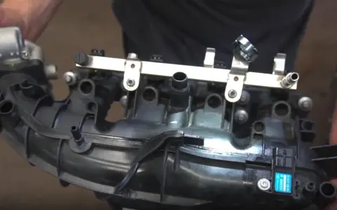Hands hold a black plastic intake manifold with a fuel rail and sensors. This part is crucial for intake manifold runner control.