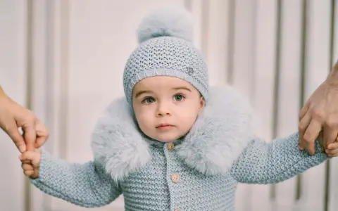 Un tierno bebé con un conjunto de punto azul claro, gorro y abrigo con cuello de pelo. Sus manos son sujetadas por adultos, como si estuviera aprendiendo a caminar. Este patrón de pelele para bebé es adorable.