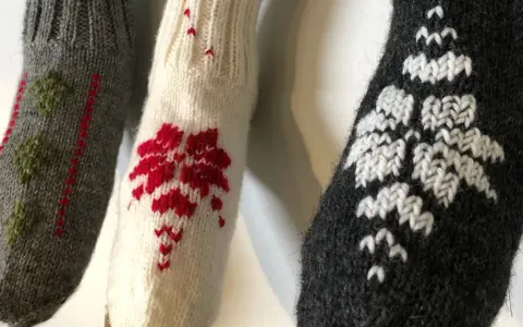 Tre stickade ärmar med mönster i maskstygn. En grå med gröna träd, en vit med röda blommor och en mörkgrå med vita snöflingor.