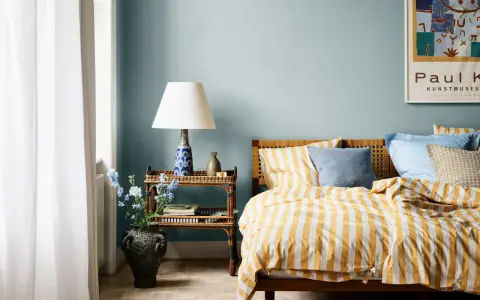 Dormitorio con paredes azul claro y ropa de cama a rayas amarillas y blancas. Los relajantes colores para dormitorios se complementan con una mesita de noche de mimbre y un jarrón con flores.