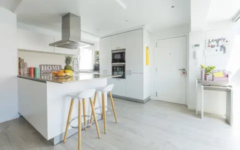 Moderna cocina blanca y gris claro con isla central, taburetes y frutas frescas.