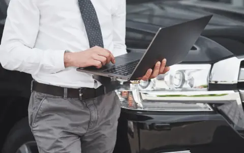 Biznesmen sprawdza na laptopie, ile czasu na przerejestrowanie auta. Elegancki mężczyzna w garniturze przy luksusowym samochodzie.