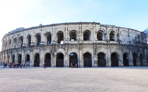 The ancient Roman amphitheater in Nîmes, France, stands majestically under a clear blue sky. Tourists gather in the plaza, ready to explore this historic marvel.
