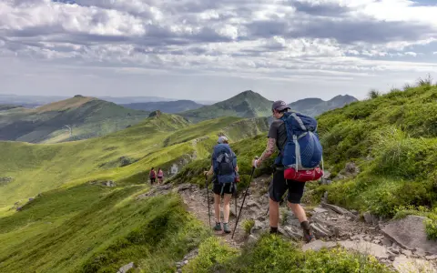 Randonnée sur le Puy Mary, des marcheurs explorent un sentier rocailleux bordé de verdure, avec des montagnes ondulantes en arrière-plan sous un ciel nuageux.
