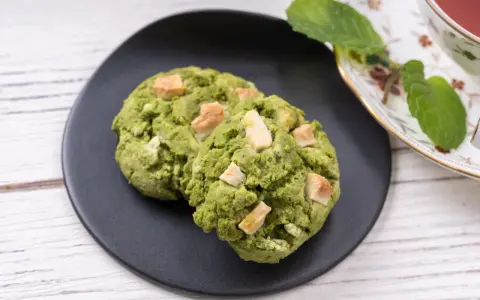 Deux cookies clairs au matcha, parsemés de morceaux de chocolat blanc, sur une assiette noire, à côté d'une tasse de thé et de feuilles de menthe.