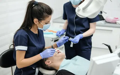 Lekarz stomatolog w masce i rękawiczkach wykonuje zabieg leczenia zębów pod narkozą. Pacjent leży na fotelu dentystycznym.