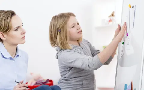 Terapeutaka z piórem w dłoni obserwuje dziewczynkę przyczepiającą rysunki do tablicy, analizując jej słabe strony.