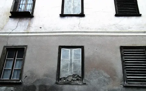 Stara elewacja z widocznymi pęknięciami i zaciekami. Okna z żaluzjami i jedno wybite.