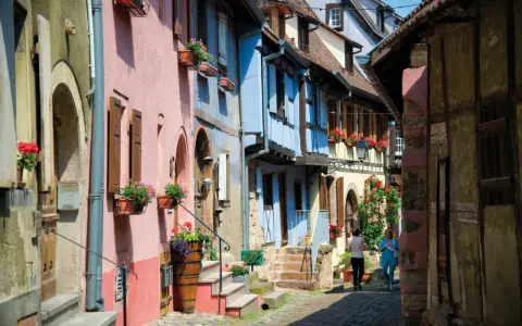Charming street with colorful houses, perfect for day trips from Strasbourg. Two women stroll past flower-filled window boxes.