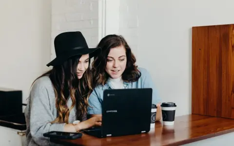Dwie młode kobiety pracują na laptopie, jedna w kapeluszu, druga w dżinsowej koszuli. Ich wspólny projekt to passe recent.