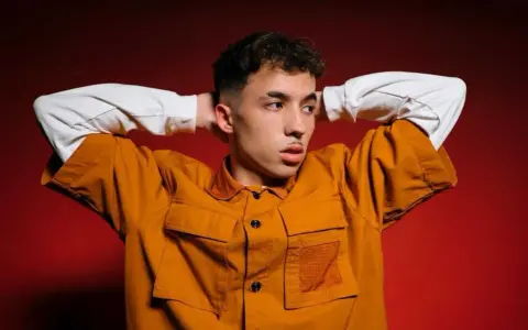 Joven con las manos detrás de la cabeza, vistiendo una chaqueta naranja y una camiseta blanca. Parece un pole cantante listo para actuar.