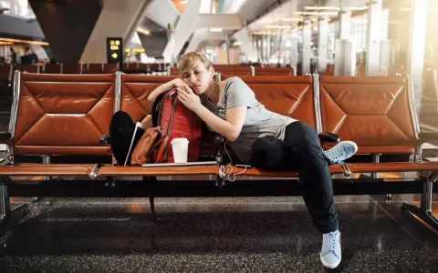 Zmęczona podróżniczka śpi na lotnisku, próbując pokonać jetlag. Obok niej plecak i kubek kawy.
