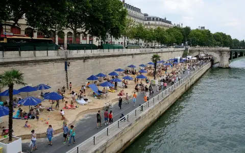 Menschen entspannen sich am **Paris strand** unter blauen Sonnenschirmen. Palmen und Gebäude säumen die Uferpromenade.