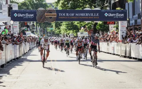 Cyclists race towards the finish line at the Tour of Somerville, America's longest-running bike race, with cheering crowds lining the street.