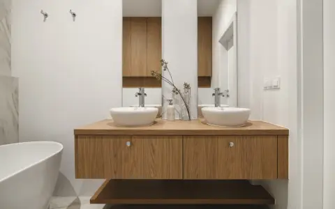Nowoczesna łazienka z dwoma umywalkami na drewnianej szafce. Minimalistyczny design, białe ceramika i ciepłe drewno tworzą harmonijną przestrzeń.