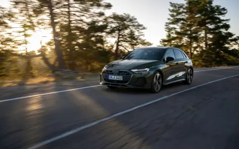 Un'auto verde scuro, un segmento premium, sfreccia su una strada panoramica al tramonto, con alberi sfocati sullo sfondo.