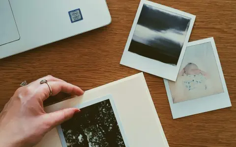 Dłoń układa zdjęcie na albumie, obok inne fotografie i laptop. Idealne miejsce, gdzie przechowywać zdjęcia z telefonu.