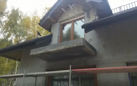 Niedokończona elewacja domu z balkonem i oknem. Na dachu siedzi ptak.