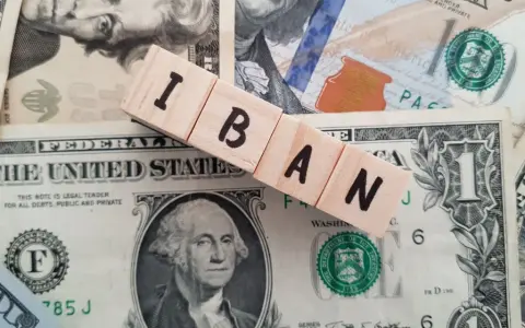 Drewniane klocki z literami IBAN leżą na banknotach dolarowych. Ile cyfr ma numer konta IBAN? To zależy od kraju, ale zazwyczaj jest to 20-34 znaki.
