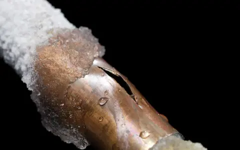 A copper pipe, partially covered in ice, shows a split from a frozen pipe repair. Water droplets cling to the damaged metal.