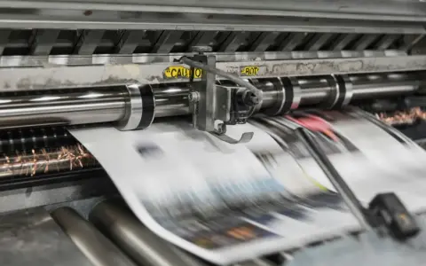Maszyna drukarska w ruchu, tworząca druk reklamowy inaczej. Papier przesuwa się szybko, ukazując kolory i kształty.