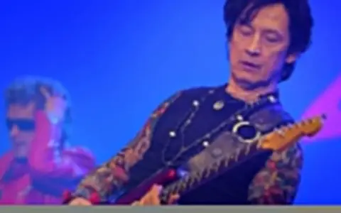 Jan Borysewicz gra na czerwonej gitarze elektrycznej, jego rękawy zdobią tatuaże.