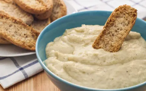 Un trozo de pan crujiente se sumerge en un cremoso baba ganoush en un cuenco azul. A un lado, hay una pila de pan tostado.