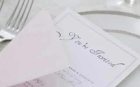 Eleganckie zaproszenie z osobą towarzyszącą, "You're Invited", na różowej kopercie, obok sztućców i talerza.