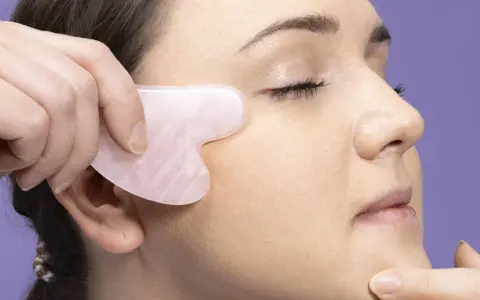 Eine Hand nutzt ein Gua Sha Stein für eine Gesichtsmassage. Das Werkzeug ist herzförmig und aus rosafarbenem Stein.