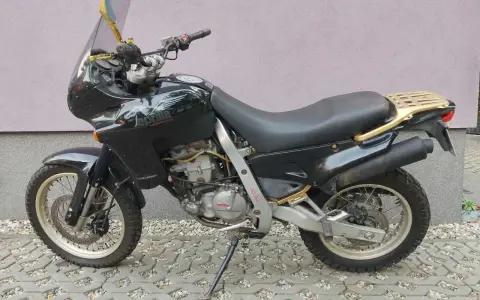 Czarny motocykl Aprilia Pegaso 650 z złotym bagażnikiem i lusterkami, zaparkowany na kostce brukowej.