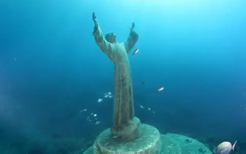 Il Cristo degli Abissi, una statua sommersa, veglia sul fondale blu, circondato da banchi di pesci che nuotano sereni.