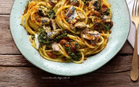 Un piatto di pasta con le sarde, condita con erbe aromatiche, pinoli e pangrattato tostato, servita su un piatto azzurro.