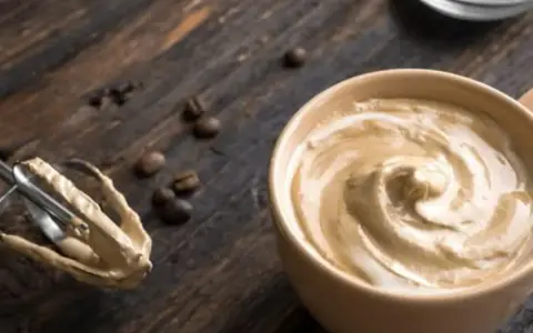 Cremige Crema al Caffè in einer Tasse, daneben ein Handmixer mit Teigresten und Kaffeebohnen auf Holz.