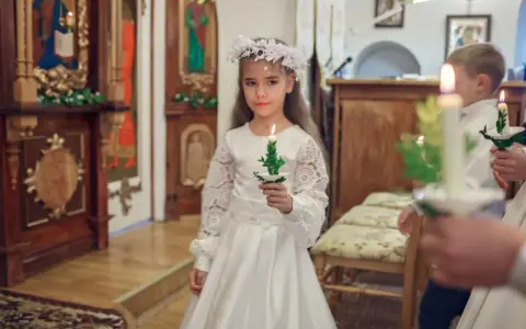 Dziewczynka w białej sukni i wianku trzyma zapaloną świecę. To ważny dzień, w którym klasa przystępuje do komunii.