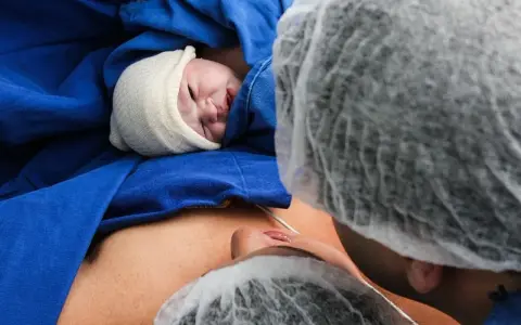 Noworodek w czepku, otulony niebieskim materiałem, leży na brzuchu mamy. Widoczna twarz dziecka i fragment twarzy mamy. To piękny moment porodu lotosowego.