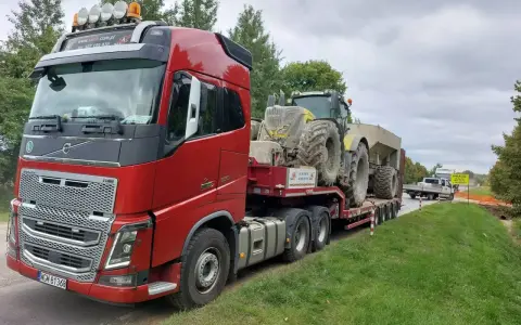 Czerwony ciągnik siodłowy przewozi koparkę na naczepie. To transport ponadgabarytowy, który wymaga specjalnych zezwoleń i przygotowania.