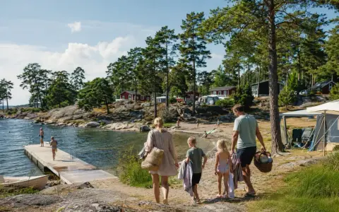 Familj på väg mot vattnet för roliga aktiviteter barn. Soligt, skärgårdsmiljö med röda stugor och camping.