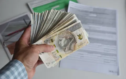 Dłoń trzyma plik banknotów 200 zł. W tle dokumenty, być może dotyczące tego, ile godzin to 0.5 etatu.
