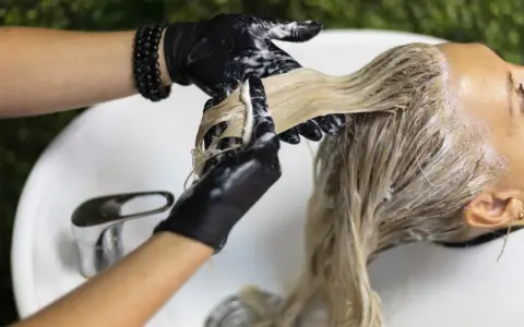 Fryzjerka w czarnych rękawiczkach nakłada odżywkę na blond włosy. Zastanawiasz się, jaki kolor na siwe włosy będzie najlepszy?