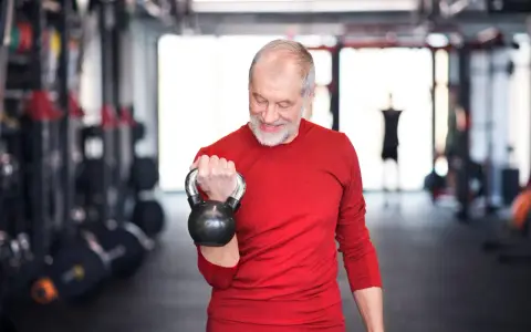 Ein älterer Mann mit Bart trainiert im Fitnessstudio mit einer Kettlebell. Ein Beispiel für Krafttraining ab 60 Jahren Trainingsplan.