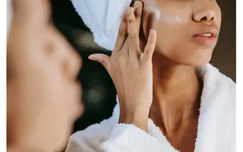 Une femme applique une crème pour l'hydratation du visage, enveloppée dans une serviette et un peignoir douillet.