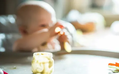 Słoiczek z puree ziemniaczanym, idealny do nauki, jak przygotować ziemniaka dla niemowlaka. W tle widać dziecko.