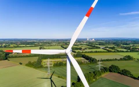 Windkraft Energie: Ein riesiger Windpark dominiert die Landschaft, umgeben von grünen Feldern und Bäumen. Im Hintergrund sind Kühltürme zu sehen.