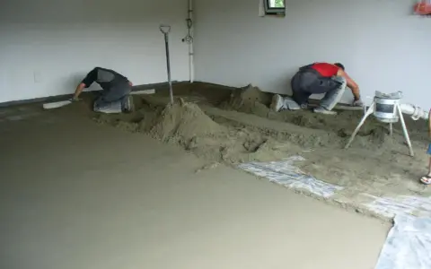 Dwóch budowlańców pracuje przy wylewce podłogi. W tle widać maszynę, która wygląda jak **mixokret**.