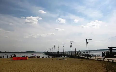 Plaża nad jeziorem Miedwie, z molo i spacerującymi ludźmi. Słoneczny dzień, chmury na niebie.