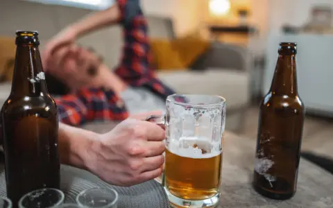 Mężczyzna leży na kanapie, trzymając kufel piwa. Wokół leżą puste butelki. Czy to oznacza, co przyspiesza spalanie alkoholu?
