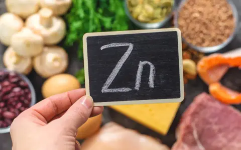 Tabliczka z napisem "Zn" na tle produktów bogatych w cynk: mięso, ryby, fasola, grzyby, jajka, ser, nasiona.