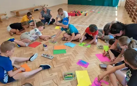 Dzieci i dorośli siedzą na podłodze, tworząc kolorowe rysunki. Wszyscy biorą udział w wesołych zabawach integracyjnych, rozwijając kreatywność.